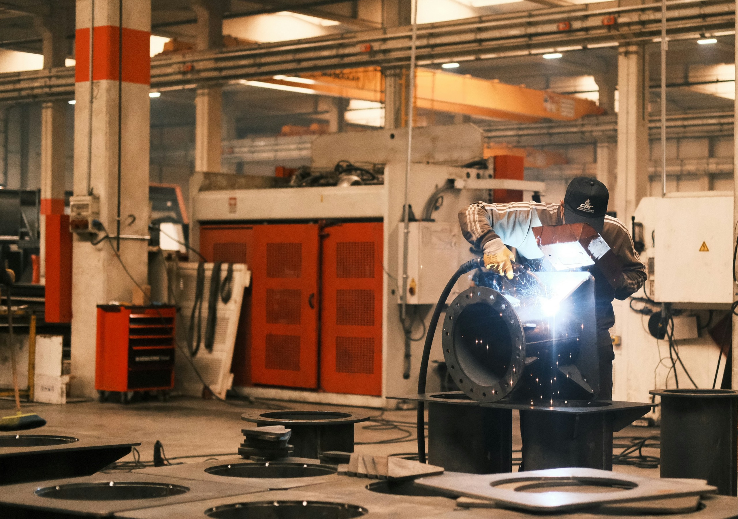 Welder joining a steel part on a busy factory floor, with sparks lighting the workpiece