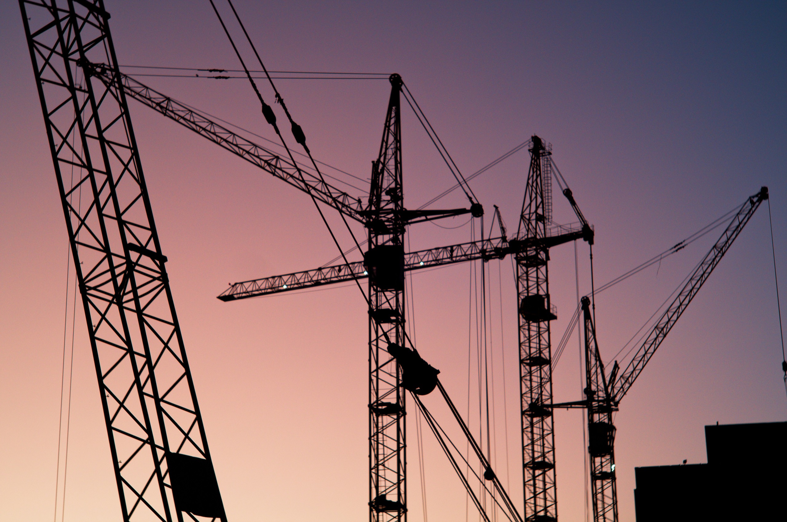 Silhouettes of tower cranes against a pink and violet evening sky